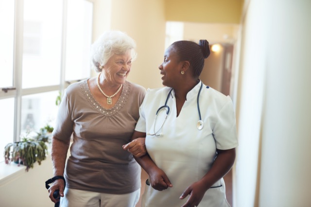 Senior woman walking in the nursing home supported by a caregiver. Nurse assisting senior woman